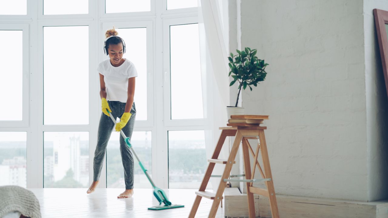 Home Young woman cleaning floor with a mop in a sunny room with large windows.