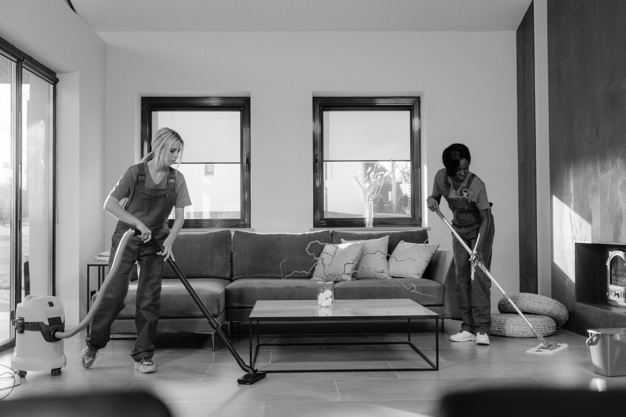 Home Two women in work clothes cleaning a modern living room using a vacuum and mop.